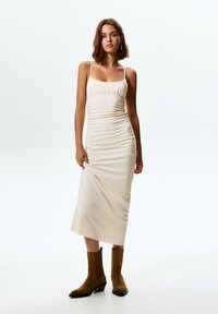 Kremno obarvana, prilegajoča midi obleka z teksturirano tkanino, nagubanjem na bodici, tankimi naramnicami in stranski razporek. Nosi se z rjavimi gležnjači.