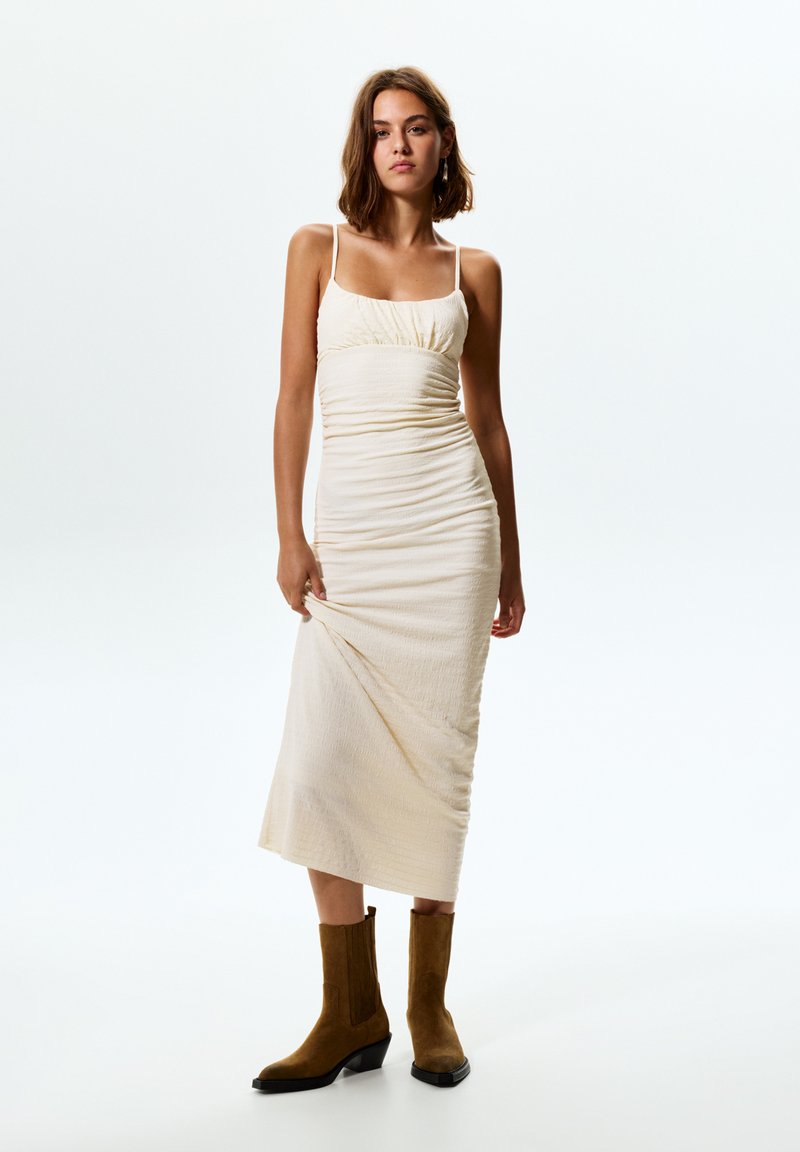 Kremno obarvana, prilegajoča midi obleka z teksturirano tkanino, nagubanjem na bodici, tankimi naramnicami in stranski razporek. Nosi se z rjavimi gležnjači.