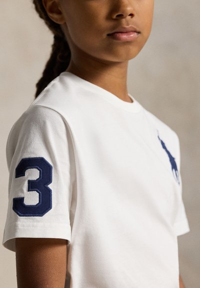 Tânăr purtând tricou alb cu mâneci scurte, cu numărul 3 bleumarin pe mânecă și sigla unui cal bleumarin pe piept, pe un fundal simplu.