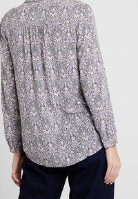 Bluzka z długim rękawem wykonana z lekkiego materiału, ozdobiona szczegółowym kwiatowym wzorem w kolorach fioletowym, beżowym i niebieskim, z marszczonym tyłem.