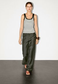 Modelen går mod kameraet iført en sort og hvid stribet tanktop, løse sorte bukser og lyserøde flip-flops. Neutral baggrund.