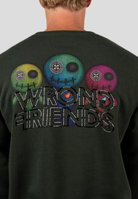 Sudadera verde caqui con gráficos de calaveras texturizados y multicolores, y el texto "WRONG FRIENDS" en letras negritas y negras en la parte posterior.