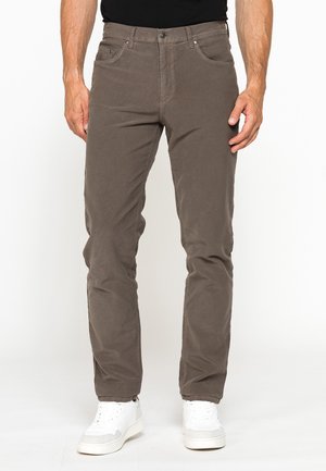 Uomo che indossa pantaloni casual marroni e scarpe da ginnastica bianche, in piedi davanti a uno sfondo chiaro uniforme con le mani rilassate lungo i fianchi.