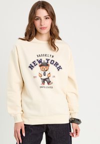 Sweatshirt crème avec un graphique d'ours portant une casquette, avec le texte "BROOKLYN NEW-YORK" et "EST. 1986" en dessous, associé à un pantalon en jean noir.