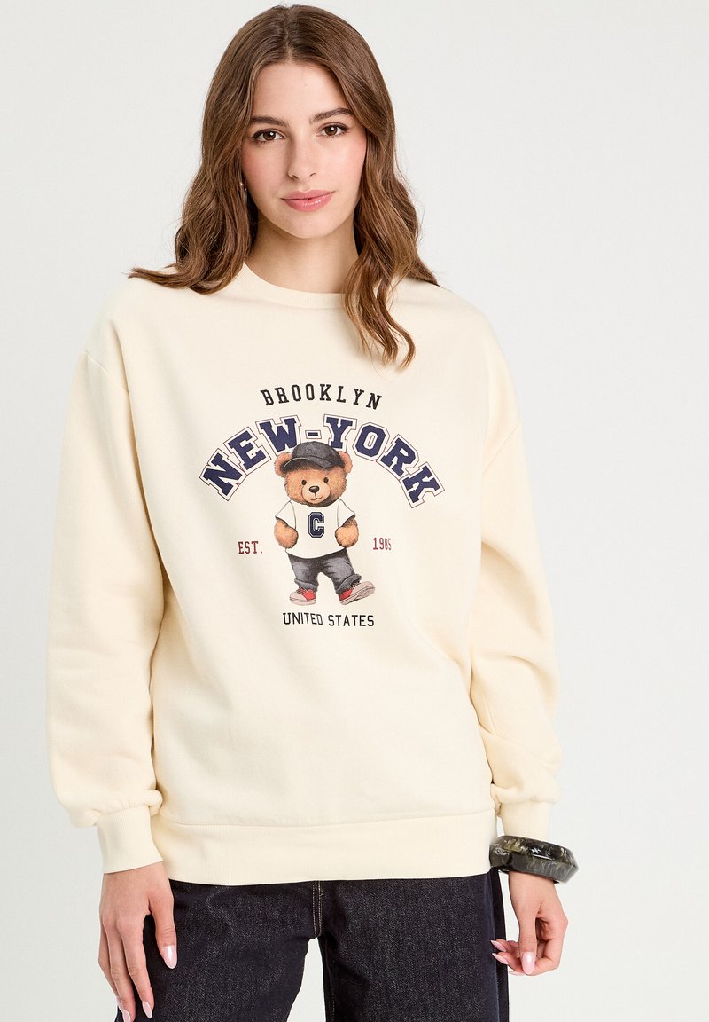 Sweatshirt crème avec un graphique d'ours portant une casquette, avec le texte "BROOKLYN NEW-YORK" et "EST. 1986" en dessous, associé à un pantalon en jean noir.