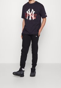 Marinblå bomulls-t-shirt med vitt och rött Yankees-logga, ihop med svarta sweatpants och svarta träningsskor med texturerad yta och snören.