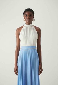 Halter-neck top i hvidt stof, parret med en plisseret blå nederdel. Nederdelen har en tynd metallisk accent i taljen. Glat tekstur.