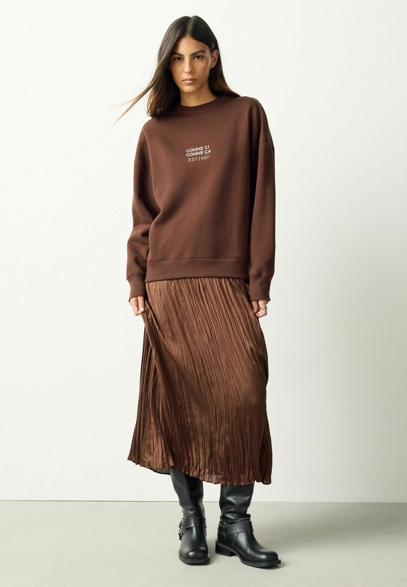 Frau trägt einen braunen Pullover mit Text, eine plissierte braune Midirock und schwarze kniehohe Stiefel, steht vor einem schlichten hellen Hintergrund.