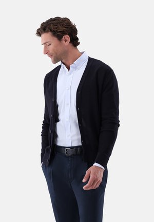 Mann mit lockigem braunem Haar, der eine schwarze, geknöpfte Strickjacke, ein weißes Hemd mit Kragen, einen dunklen Gürtel und marineblaue Hosen trägt, schaut nach unten und hat die Hände in den Taschen.