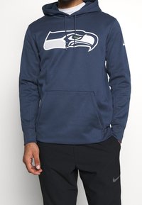 Felpa con cappuccio di colore blu navy realizzata in tessuto con una texture liscia, caratterizzata da un grande logo dei Seattle Seahawks in bianco e verde, con una tasca frontale.