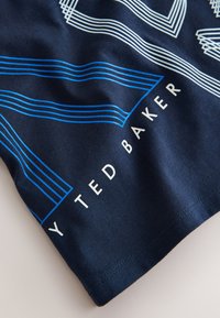 Laivastonsininen kangas, jossa on valkoisia ja sinisiä ääriviivoin kuvioituja geometrisia muotoja ja valkoinen teksti "TED BAKER" pohjareunaa pitkin.