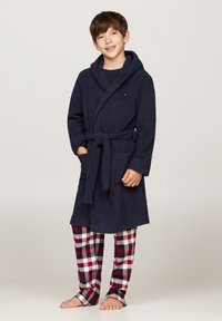 Navyblauwe fleece badjas met een capuchon en riem, voorzien van twee voorzakken. Rood-witte geruite pyjamabroek maakt de outfit compleet.
