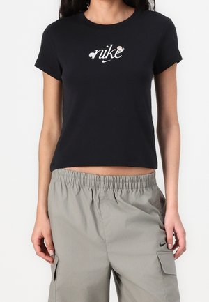 Kobieta ubrana w czarny krótki crop top Nike z kwiatowym logo oraz szare spodnie cargo z logo Nike na kieszeni.