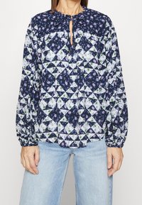 Langsleeve blouse met een marineblauwe bloemenpatchwork ontwerp met vierkante en driehoekige vormen, een keyhole hals en elastische manchetten.