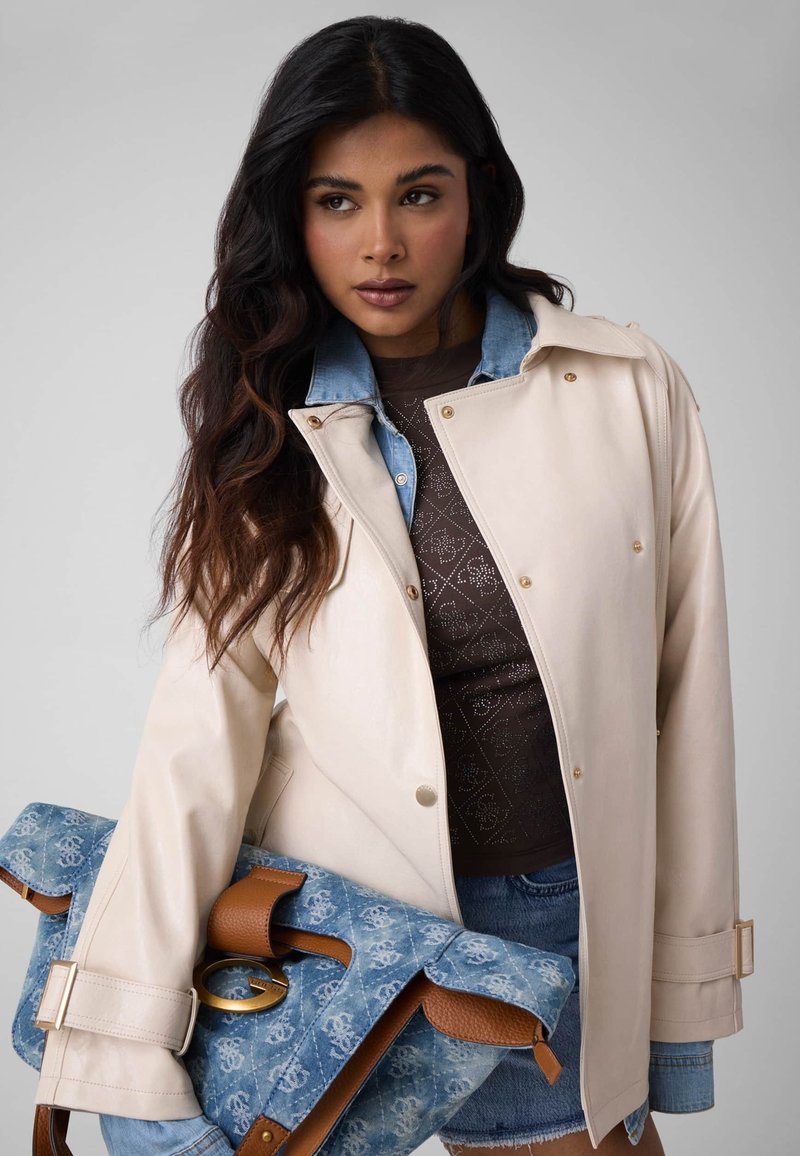 Femme aux longs cheveux foncés, portant une veste beige et une chemise en denim, tenant un grand sac à main bleu à motifs avec des touches de cuir marron.