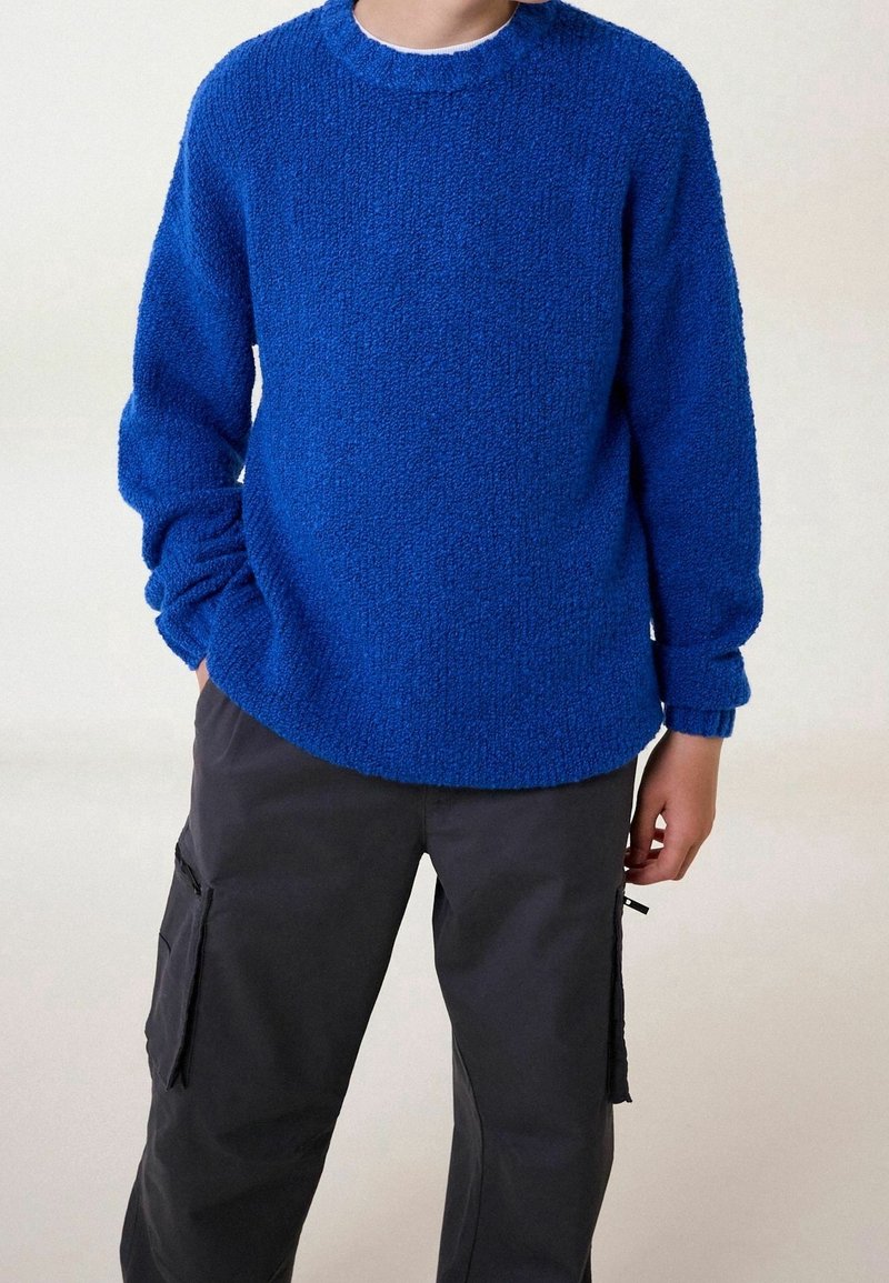 Personne portant un pull bleu texturé à manches longues et un pantalon cargo gris foncé, debout avec une main dans la poche devant un fond uni.