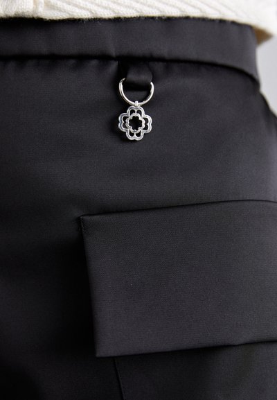 Μαύρο παντελόνι από ύφασμα με μπροστινή τσέπη και διακοσμητικό ασημένιο λουλουδένιο γούρι προσαρτημένο σε θηλιά. Λείο ύφασμα, εφαρμοστή γραμμή.