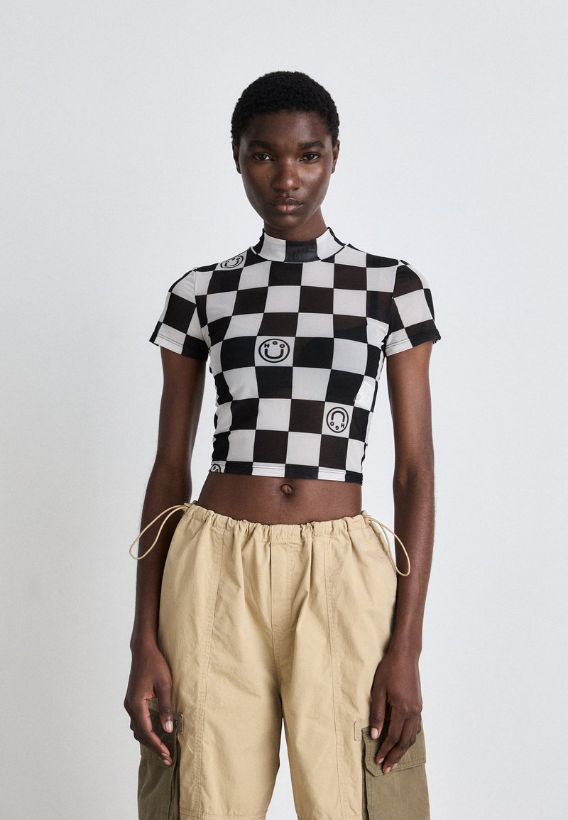 Crno-bijeli karirani crop top s grafikom s osmijehom, s visokim ovratnikom i kratkim rukavima. Nosite ga uz bež cargo hlače.