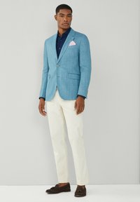 Blazer in lino azzurro chiaro con revers a scultura, due tasche frontali e un fazzoletto da taschino rosa, abbinato a pantaloni crema e mocassini scuri.