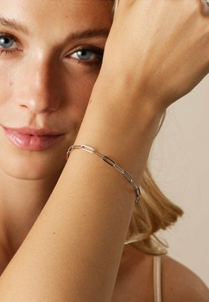 Gros plan d'une femme à la peau claire et aux yeux bleus portant un délicat bracelet en argent à maillons sur son poignet levé.