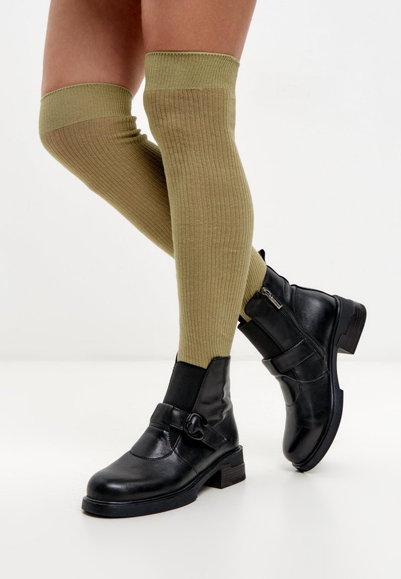 Botas de tobillo de cuero negro con un acabado texturizado, detalle de hebilla metálica y un ligero tacón, combinadas con calcetas de canalé en color verde oliva que llegan por encima de la rodilla.