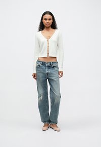 Donna con capelli ricci che indossa un cardigan bianco a maniche corte con ganci, jeans blu larghi e strappati, e scarpe basse beige, in piedi dritta.