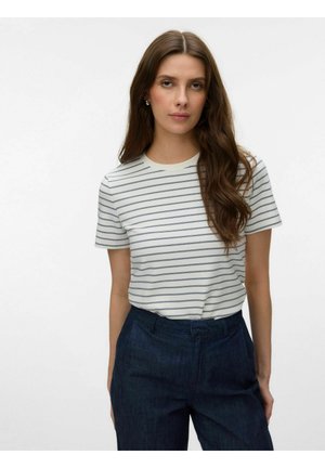 Donna con lunghi capelli castani che indossa una maglietta bianca a maniche corte con sottili strisce orizzontali nere e pantaloni blu scuro a vita alta.