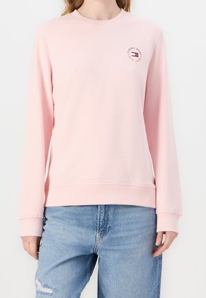 Femme portant un sweat-shirt à manches longues rose clair avec un petit logo rond Tommy Jeans sur la poitrine et un jean bleu clair déchiré.
