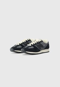 Black sneakers with a suede and glossy material mix, cream laces, and a rubber sole with a textured grip pattern. Classic design elements.
