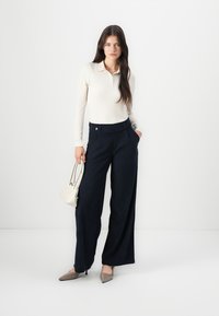 Polo shirt bianco a maniche lunghe a coste, pantaloni a gamba larga navy a vita alta con bottoni frontali e una piccola borsa color crema. Scarpe a punta in tonalità neutre.