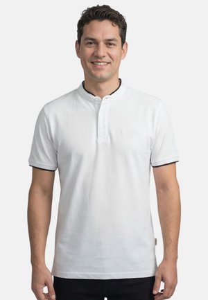 Homme souriant aux cheveux courts et bouclés, portant un polo blanc à manches courtes avec un liseré noir, debout devant un fond uni.