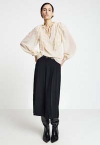 Cremefarbene Bluse mit Spitzenverzierungen, gerafften Ärmeln, tiefem V-Ausschnitt, kombiniert mit schwarzen weit geschnittenen Hosen und glänzenden schwarzen kniehohen Stiefeln.