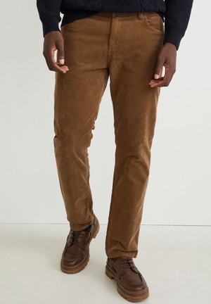 Mężczyzna w brązowych spodniach sztruksowych i ciemnobrązowych skórzanych butach, stojący na tle jasnego, jednolitego tła.