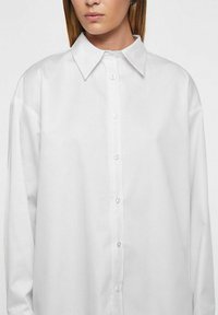 Valkoinen, pitkähihainen paita, valmistettu sileästä kankaasta. Siinä on klassinen kaulus ja edessä napitettava suljenta sekä rento, ylisuurikokoinen istuvuus.