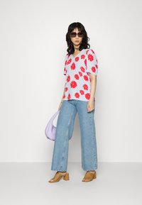 T-shirt de manga curta floral em azul claro com flores vermelhas arrojadas, combinado com jeans de lavagem clara de perna larga, mules castanhas perfuradas de salto e uma bolsa lilás.