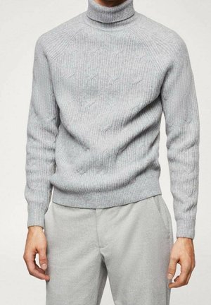 Mężczyzna ubrany w jasnoszary sweter z golfem o splocie warkoczowym oraz pasujące jasnoszare spodnie, stojący na jednolitym tle.