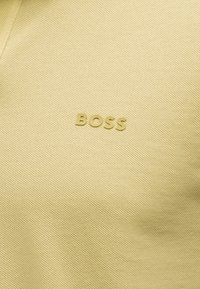 Ανοιχτό κίτρινο υφασμάτινο με υφή και ανυψωμένα γράμματα που σχηματίζουν τη λέξη "BOSS" στο κέντρο της επιφάνειας.