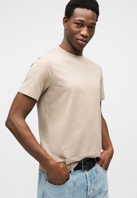 Μπεζ βαμβακερό t-shirt με κοντά μανίκια, στρογγυλή λαιμόκοψη και ελάχιστο λογότυπο. Συνδυασμένο με ανοιχτό μπλε τζιν παντελόνι και μαύρη ζώνη.