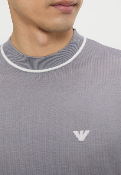 Maglioncino a maniche corte, di colore grigio chiaro, con colletto a coste bianco e un piccolo logo bianco sul petto, caratterizzato da un tessuto a maglia liscia e strutturata.