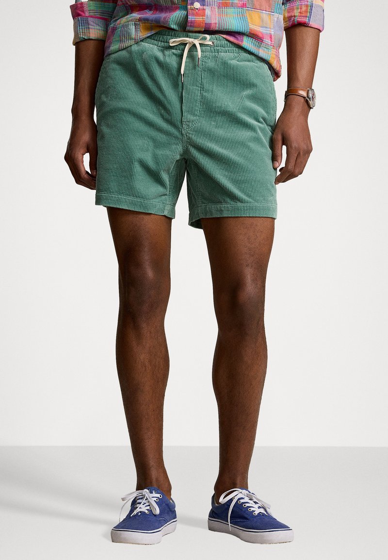 Shorts en velours côtelé vert avec une taille élastique et un cordon de serrage, présentant une surface texturée. Assortis à des baskets bleues et une chemise à carreaux.