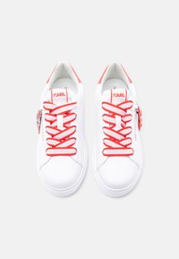 Ζευγάρι λευκά χαμηλά sneakers με κόκκινα και λευκά κορδόνια, κόκκινες ενισχύσεις στη φτέρνα και κόκκινο λογότυπο "KARL" στη γλώσσα και την πλευρά.