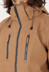 Brun huvtröja med grå dragkedjor, som har två dragkedjefickor och snörjusteringar, tillverkad av ett texturerat tyg.