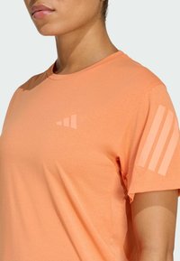 Ferskenfarget treningst-skjorte laget av lett stoff, med tre vertikale striper på øvre venstre erme og en liten logodetalj.