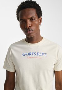 Cremefarvet t-shirt med korte ærmer, der har blå "SPORTS DEPT." tekst og rød "hummel PERFORMANCE" nedenunder. Glat stof, afslappet fit.