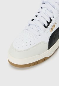 Hvit sneakers med svart stripe, hvite snøringer, perforert tå, beige mellomsåle, gummisåle og gull Puma-logo på siden.