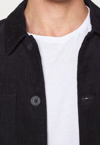 Svart kordjacka med knäppt krage, framfickor och strukturerad design, lager på en vit t-shirt.