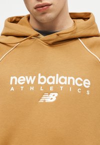 Κοντινό πλάνο ατόμου που φοράει φούτερ με κουκούλα της New Balance Athletics σε χρώμα μουσταρδί κίτρινο, με λευκό λογότυπο και κορδόνια.