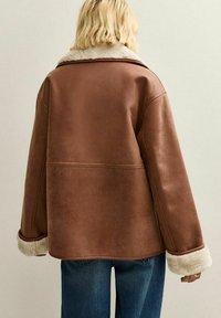 Giubbotto di pelle marrone con colletto in shearling; presenta una vestibilità oversize e maniche larghe. Texture morbida ed elementi di design senza cuciture visibili.
