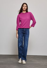 Suéter texturizado rosa con capucha y mangas de tres cuartos, combinado con jeans de pierna ancha de cintura alta azules y zapatillas blancas, de pie sobre un suelo gris.