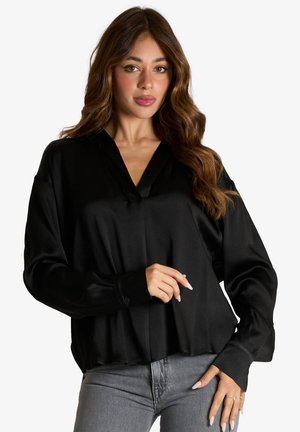 Donna con lunghi capelli mossi castani, che indossa una camicetta nera in raso e jeans grigi, in piedi davanti a uno sfondo bianco uniforme.